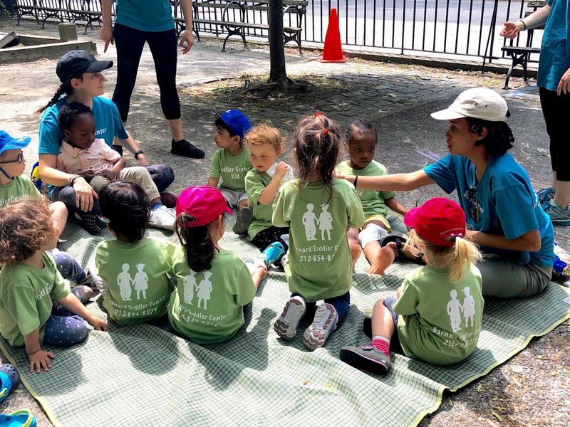 group of toddlers on a blanket in a park, with a couple of Barnard students