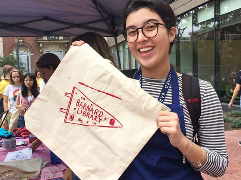 Screenprinting event. A student excitedly holds up a canvas bag with the Barnard Library pennant on it. 